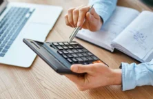 main tenant un stylo et une calculatrice, un ordanateur portable et un carnet posé sur la table(© Freepik)